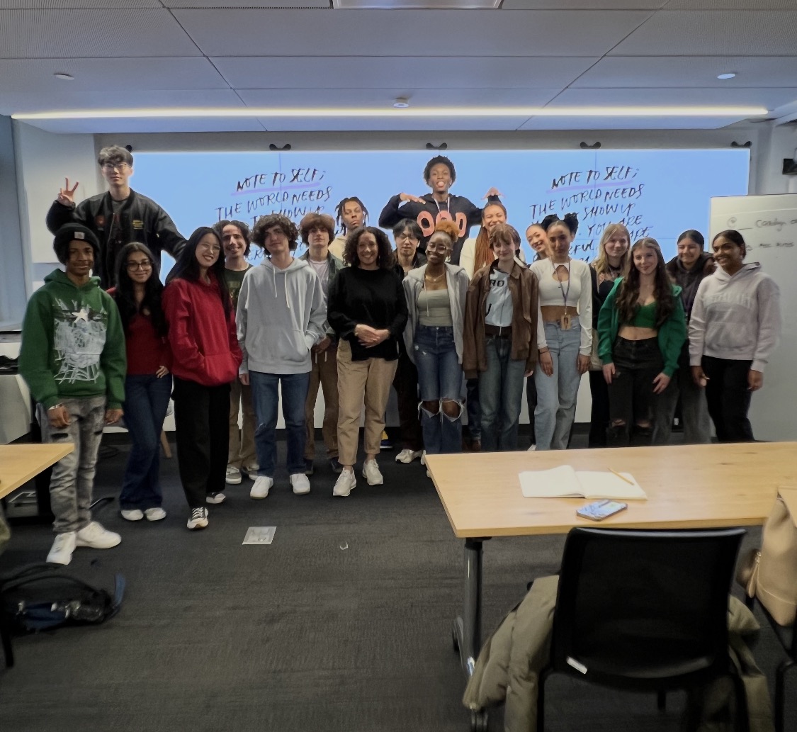 Carolyn Ortiz with a full class of students in front of a 'Note to self: the world needs you to show up' screen